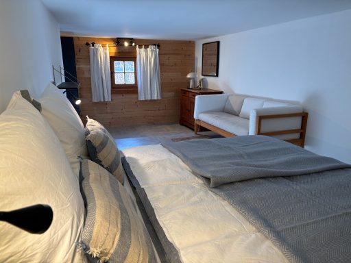 Helles Schlafzimmer mit Doppelbett, Sofaecke und Fenster mit Vorhängen.