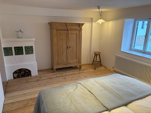 Helles Schlafzimmer mit Holzfußboden, Kachelofen, Kleiderschrank und Fenster.